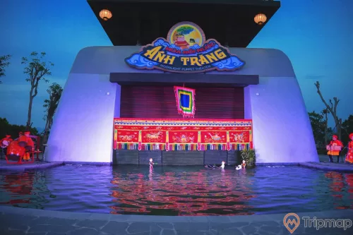Sân khấu múa rối nước Ánh Trăng, nhiều người mặc đồ đỏ đang đánh trống, nhiều hình nộm dưới nước, ảnh chụp buổi tối