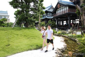 Vườn Nhật Bản Zen Garden, 2 người đang đi bộ trên nền đường màu xám, nhiều cây xanh, nhà có mái ngoái màu xanh, hồ nước nhiều viên sỏi, ảnh chụp ban ngày