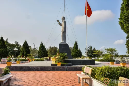 Ảnh chụp bên trái bức tượng Bác Hồ tại khu di tích lưu niệm Hồ Chủ tịch trên đảo Cô Tô, cây cối trong khuôn viên xanh tươi, cột cờ quốc kỳ Việt Nam đang bay phất phới, ghế đá đặt cạnh bồ hoa, bầu trời nhiều mây, ảnh chụp ngoài trời