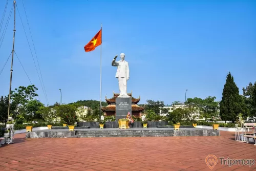 Đài tưởng niệm bác Hồ tại khu di tích lưu niệm Hồ Chủ tịch trên đảo Cô Tô, bầu trời trong xanh, bức tượng bác hồ đứng đang giơ tay màu trắng, cột cờ lá quốc kỳ Việt Nam đang bay phất phới, cây cối trong khuôn viên xanh tươi, phía sau là đền thờ bác Hồ, ảnh chụp ngoài trời