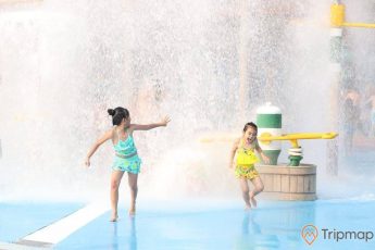 Hai đứa trẻ chơi đùa bên trò Quốc đảo kì diệu, ảnh chụp ngoài trời, một bé gái mặc trang phục xanh và một bé gái trang phục màu vàng đang chạy trong khu vui chơi, trò chơi nước